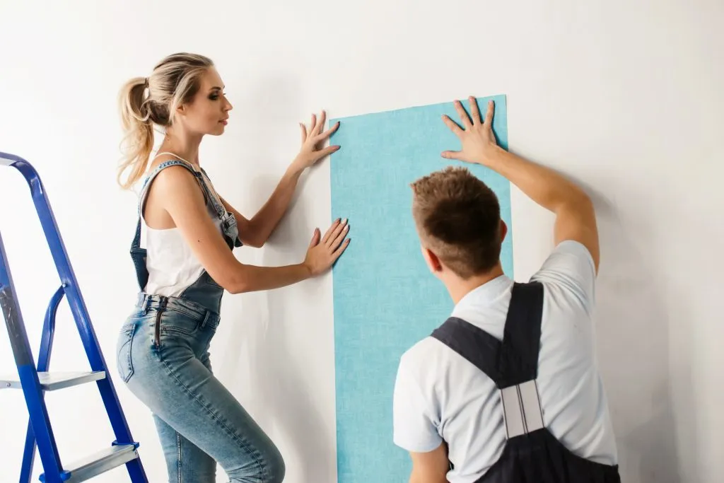 Funny couple, woman and her husband wallpapering the walls using a metal ladder. Home renovation and moving to new house concept